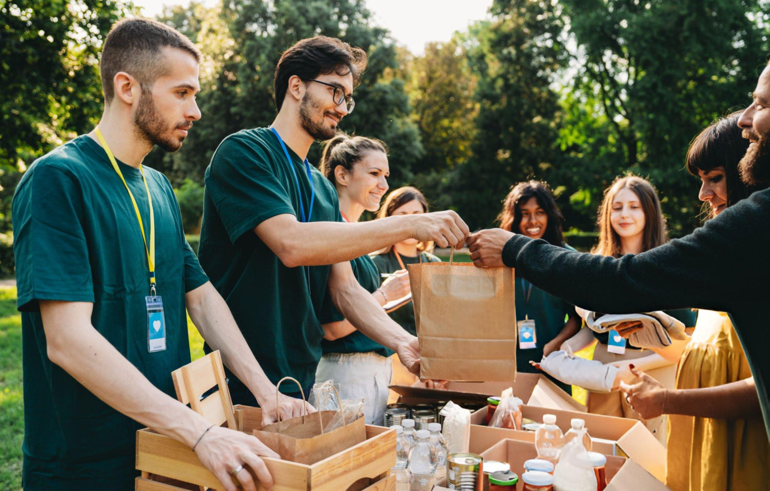 Équipe de bénévoles en tee-shirts verts distribuant des denrées alimentaires dans des sacs en carton lors d'une action solidaire en plein air.