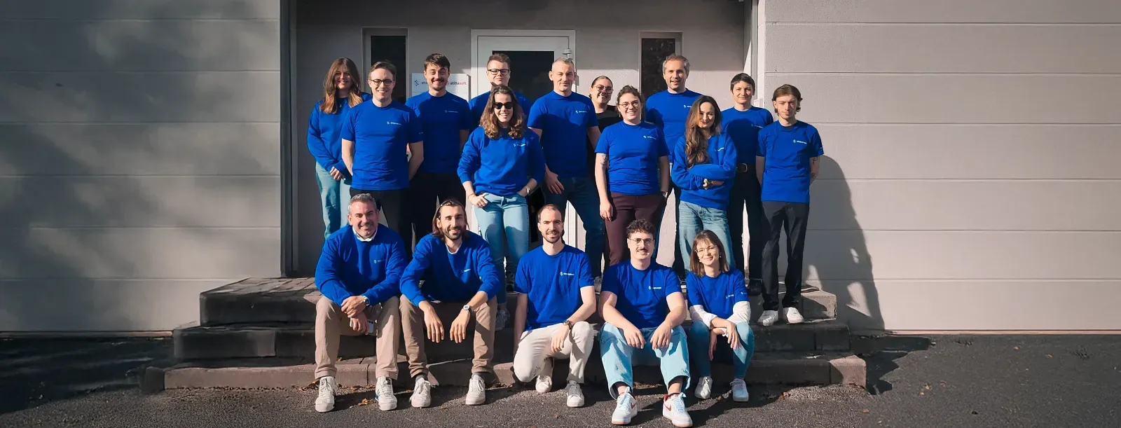 Équipe de 20 personnes en tee-shirts bleus posant devant un bâtiment blanc.