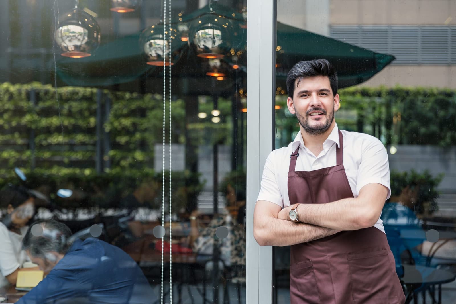Homme souriant en tablier bordeaux devant la vitrine d'un restaurant ou café.