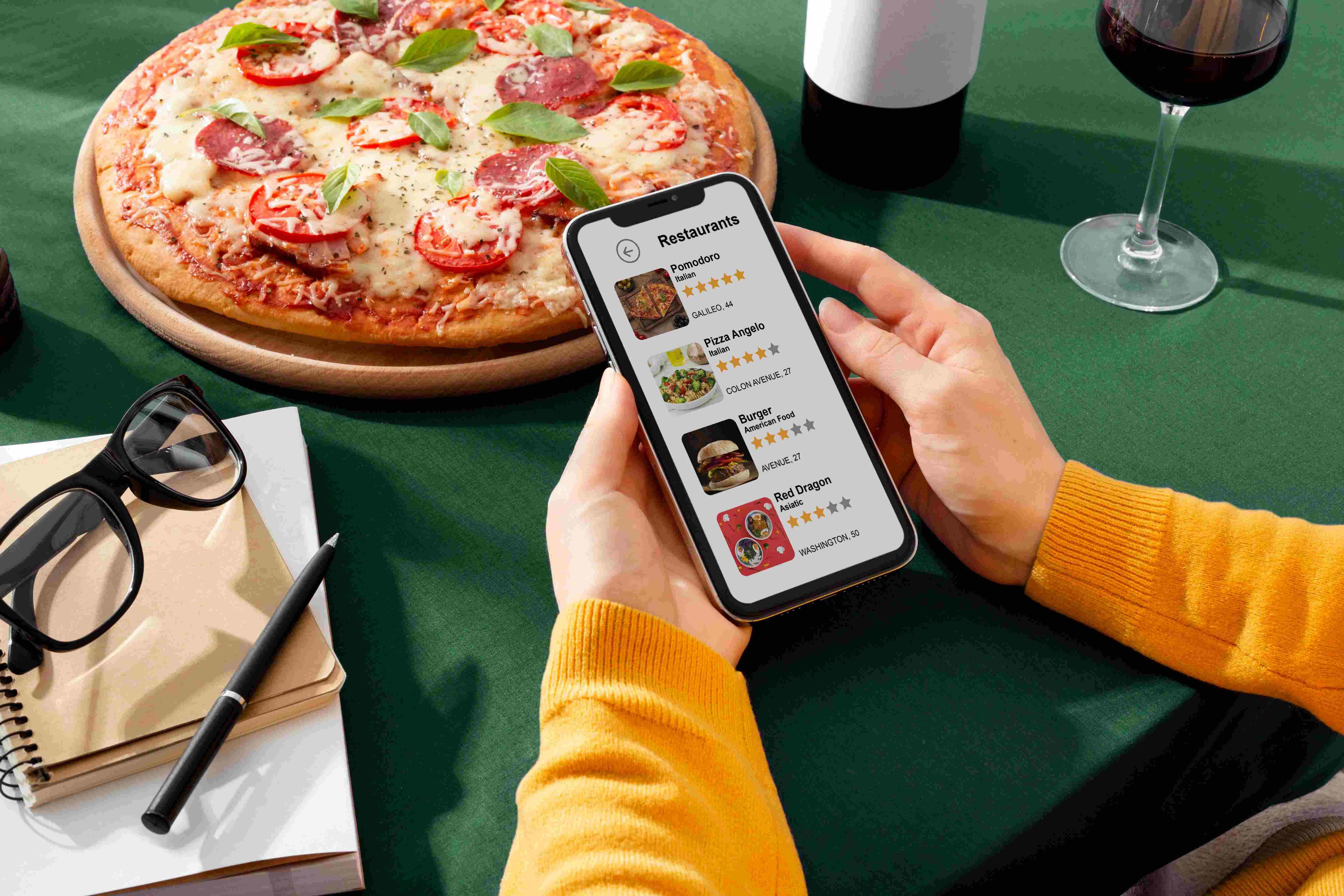 Personne consultant une application de restaurants sur smartphone à côté d'une pizza et un verre de vin.