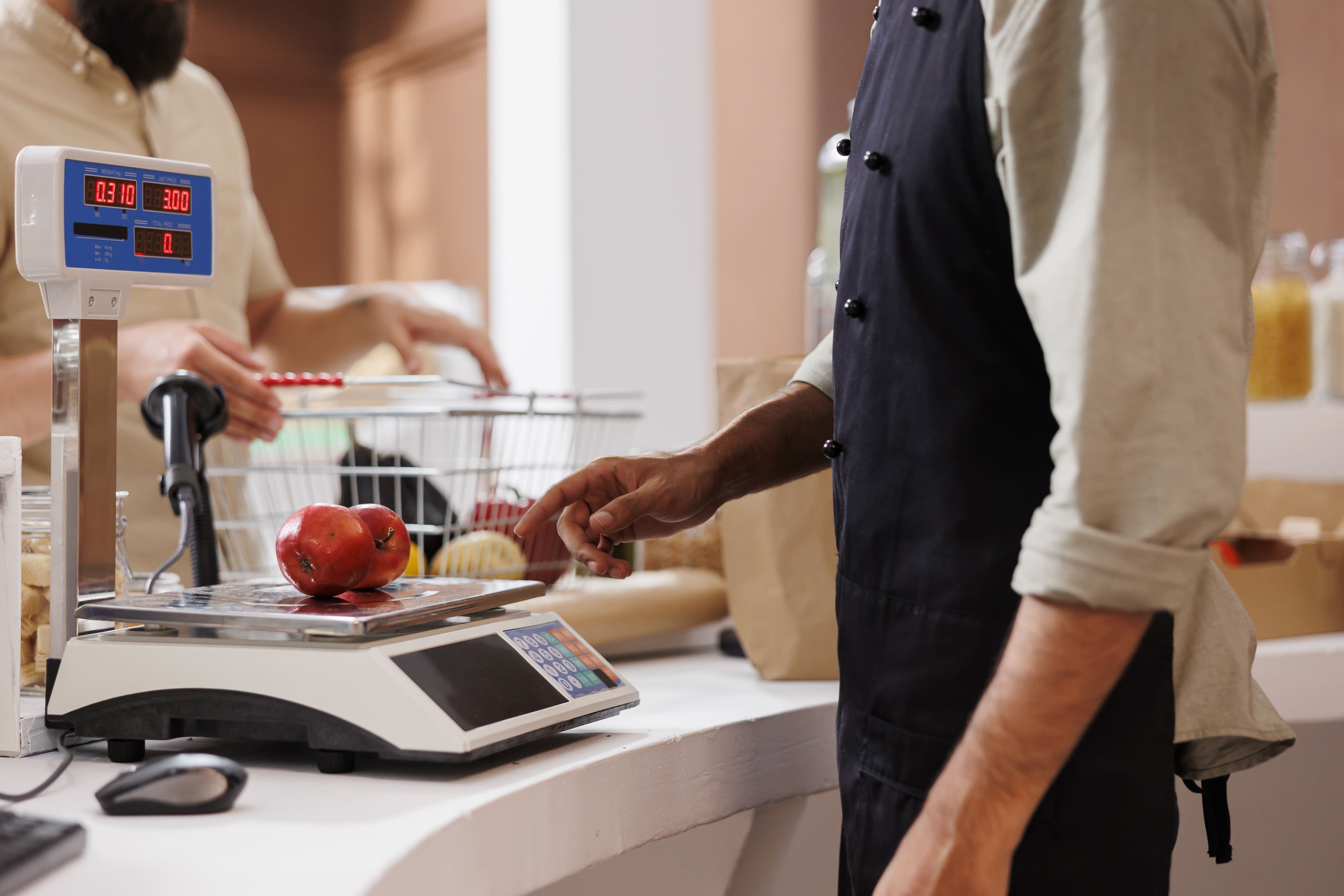 Client pesant une tomate à la caisse d'un magasin avec balance électronique affichant le prix.