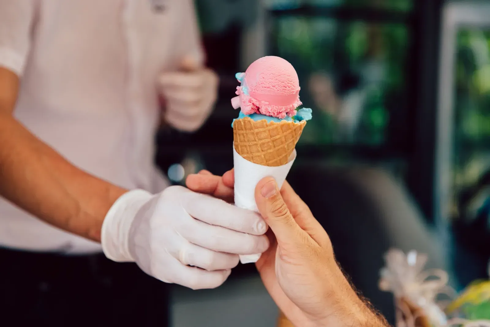 Une main reçoit un cornet de glace rose et bleu tenu par une personne en magasin.
