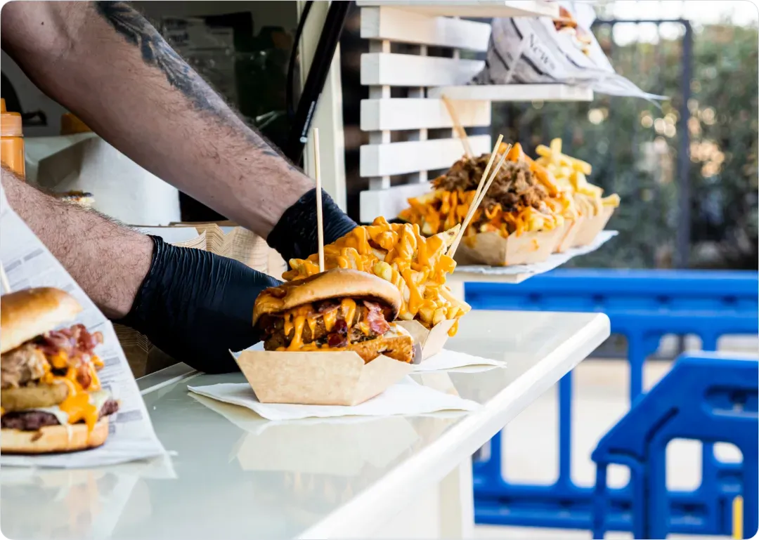 Un vendeur en gants noirs prépare des burgers garnis de fromage et frites dans un food truck en plein air.