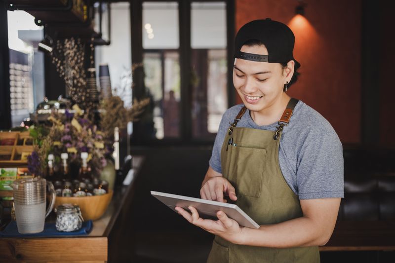 Jeune vendeur en tablier olive consulte une tablette dans un magasin avec plantes décoratives.