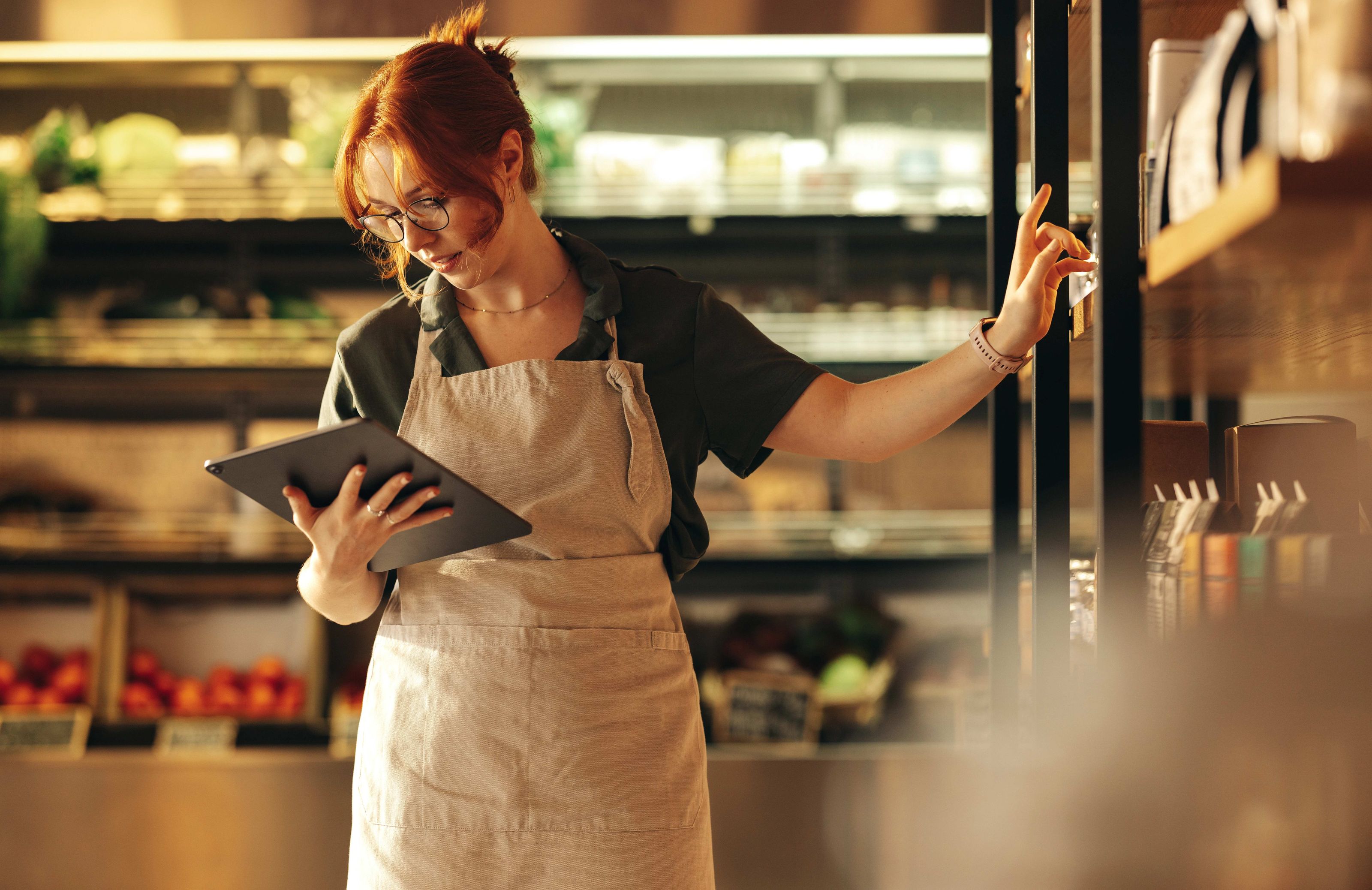 Femme avec tablette en apron gère l'inventaire devant les rayons réfrigérés d'une épicerie.