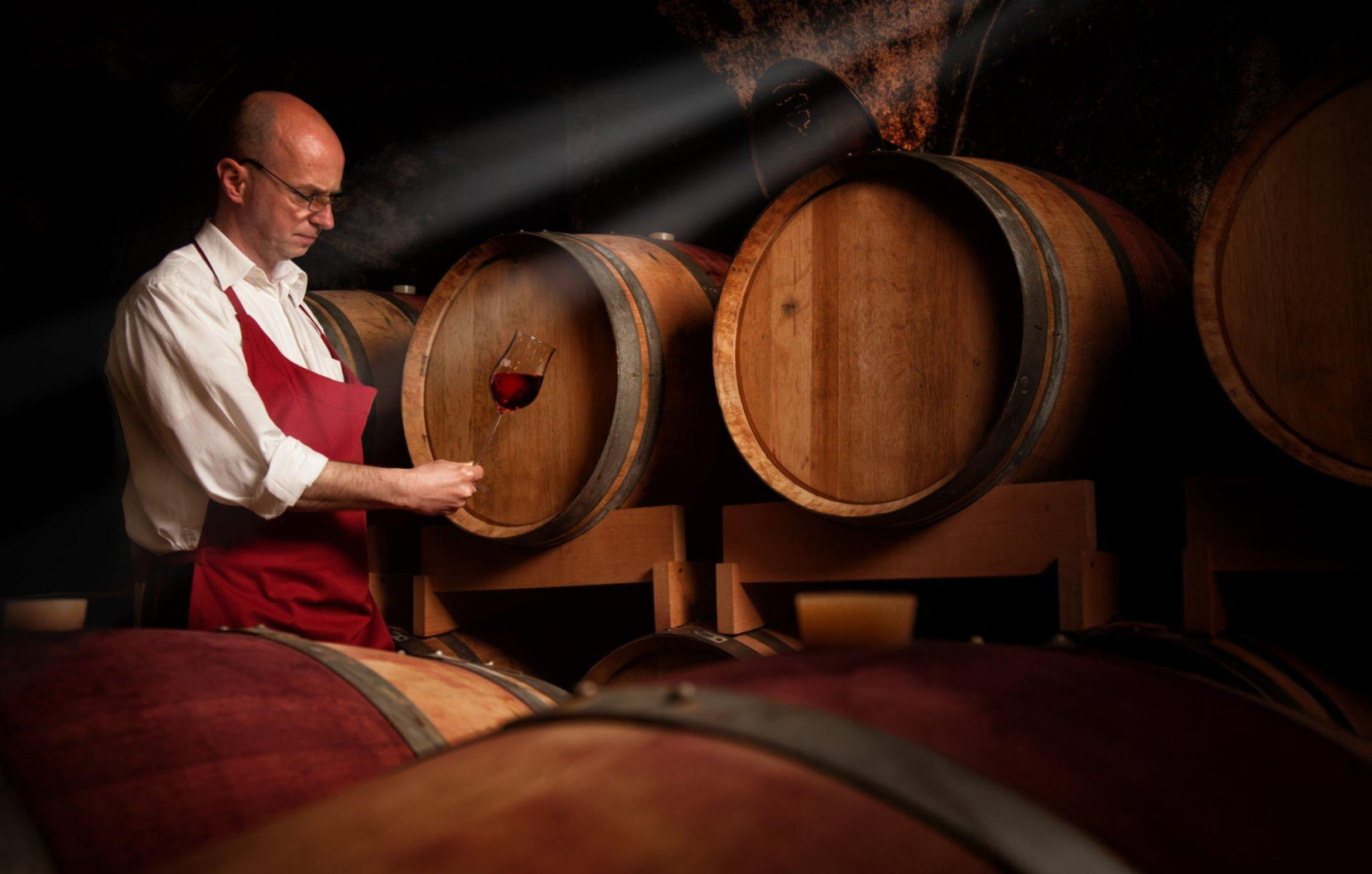 Sommelier en apron rouge contrôlant un tonneau de vin rouge en cave de vieillissement.
